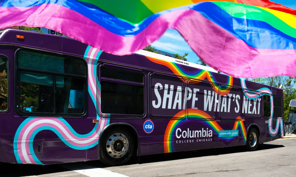 Side view of a purple CTA bus wrapped in Columbia College Chicago’s ‘Shape What’s Next’ Pride design, with rainbow and trans flag stripes, under a waving rainbow flag