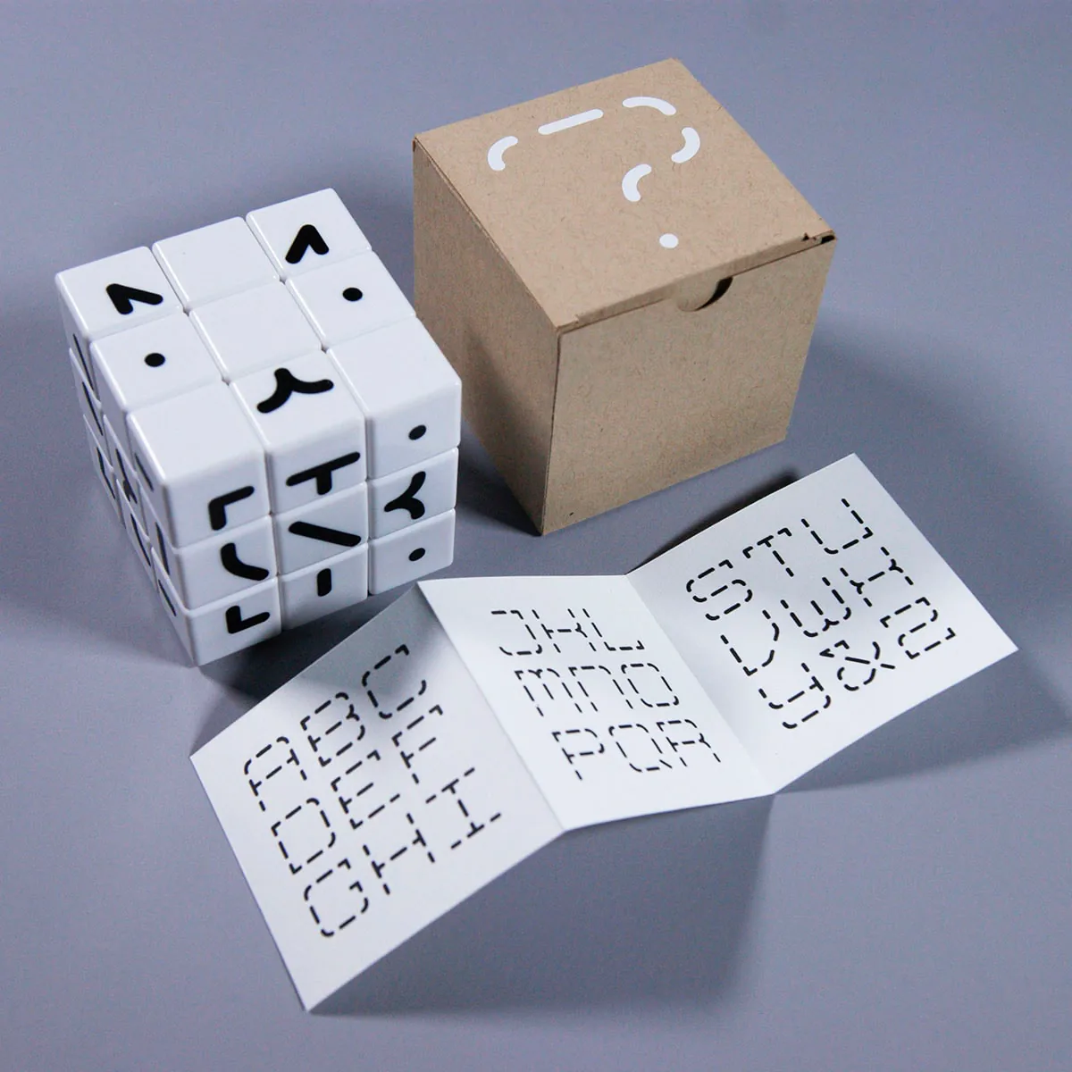 Erno Type puzzle cube next to its kraft packaging box, printed with a question mark, and an unfolded instruction booklet showing the alphabet key