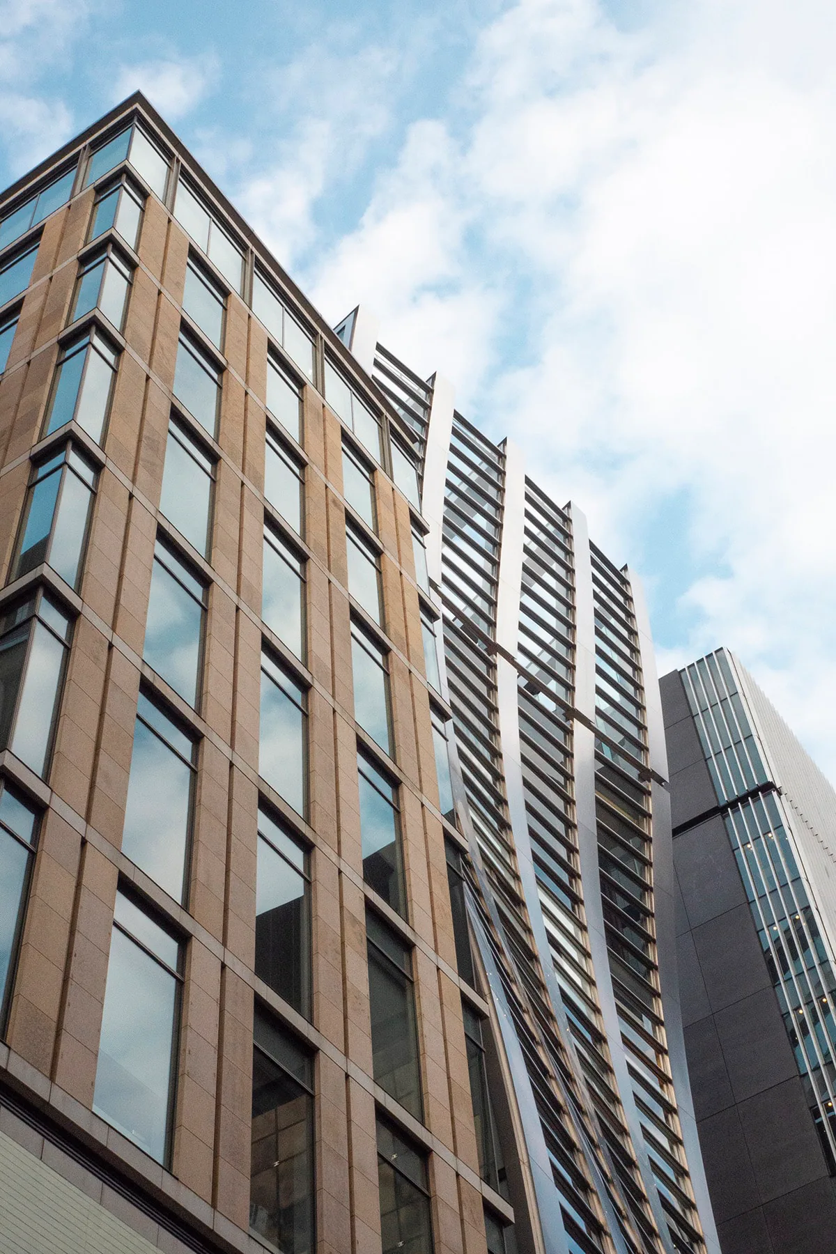 Looking straight up at an angular stone-and-glass building with slatted metal fins against a cloudy sky