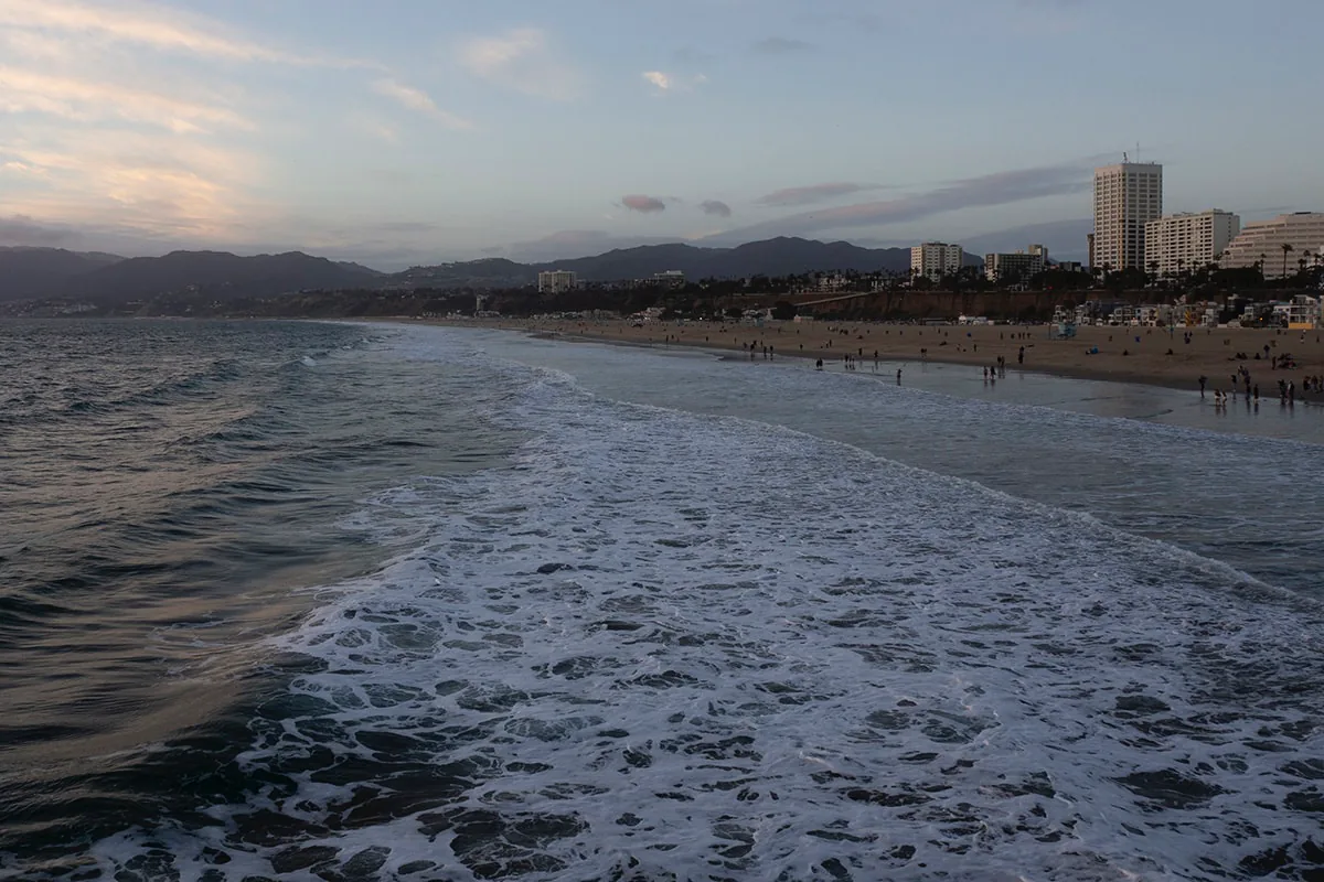 Waves roll toward a sandy beach at dusk with a distant skyline and hills fading into the haze