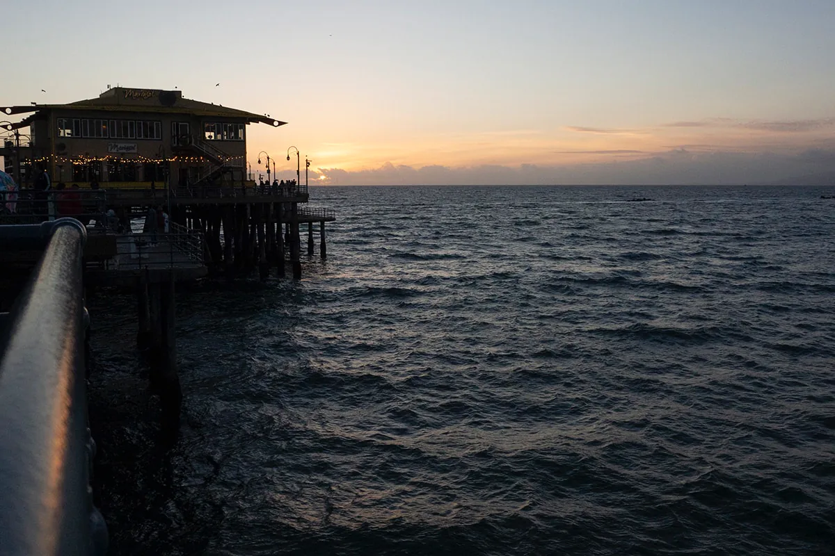 Silhouetted pier building glows with string lights as the sun sets over a calm ocean