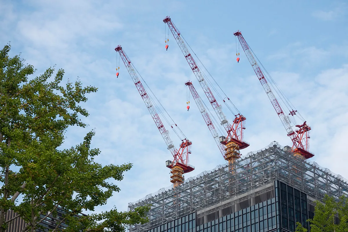 Three red tower cranes rise above a mesh-wrapped high-rise under construction against a bright blue sky