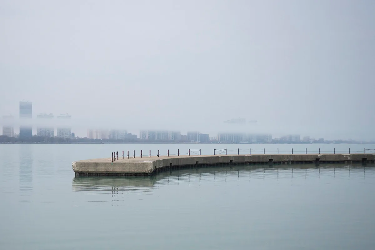 Curved concrete pier stretches into calm water toward a fog-shrouded city skyline