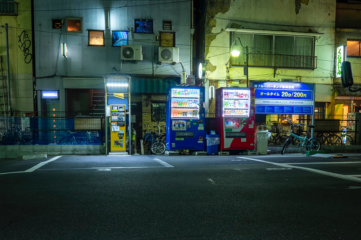 Glowing blue and red vending machines light up a quiet nighttime street corner with parked bicycles