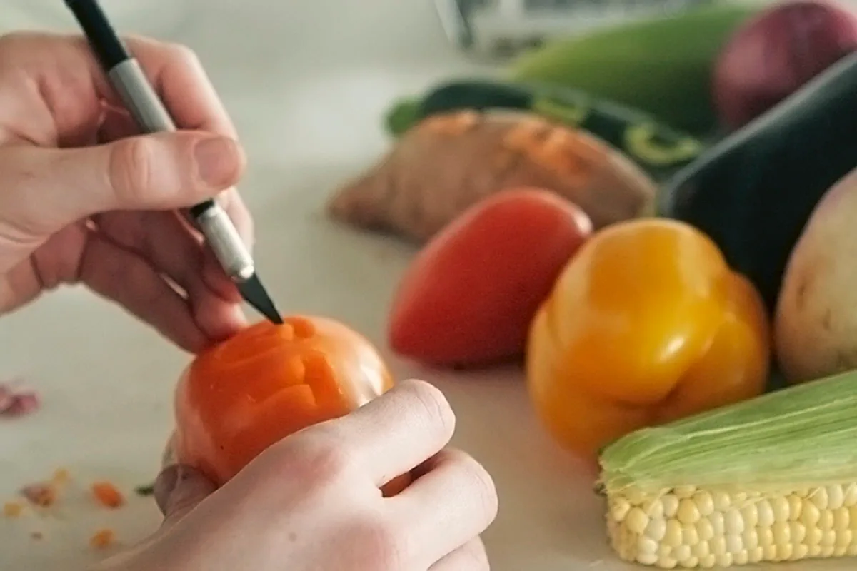 Process photo of a hand engraving a letterform into a tomato with a carving tool