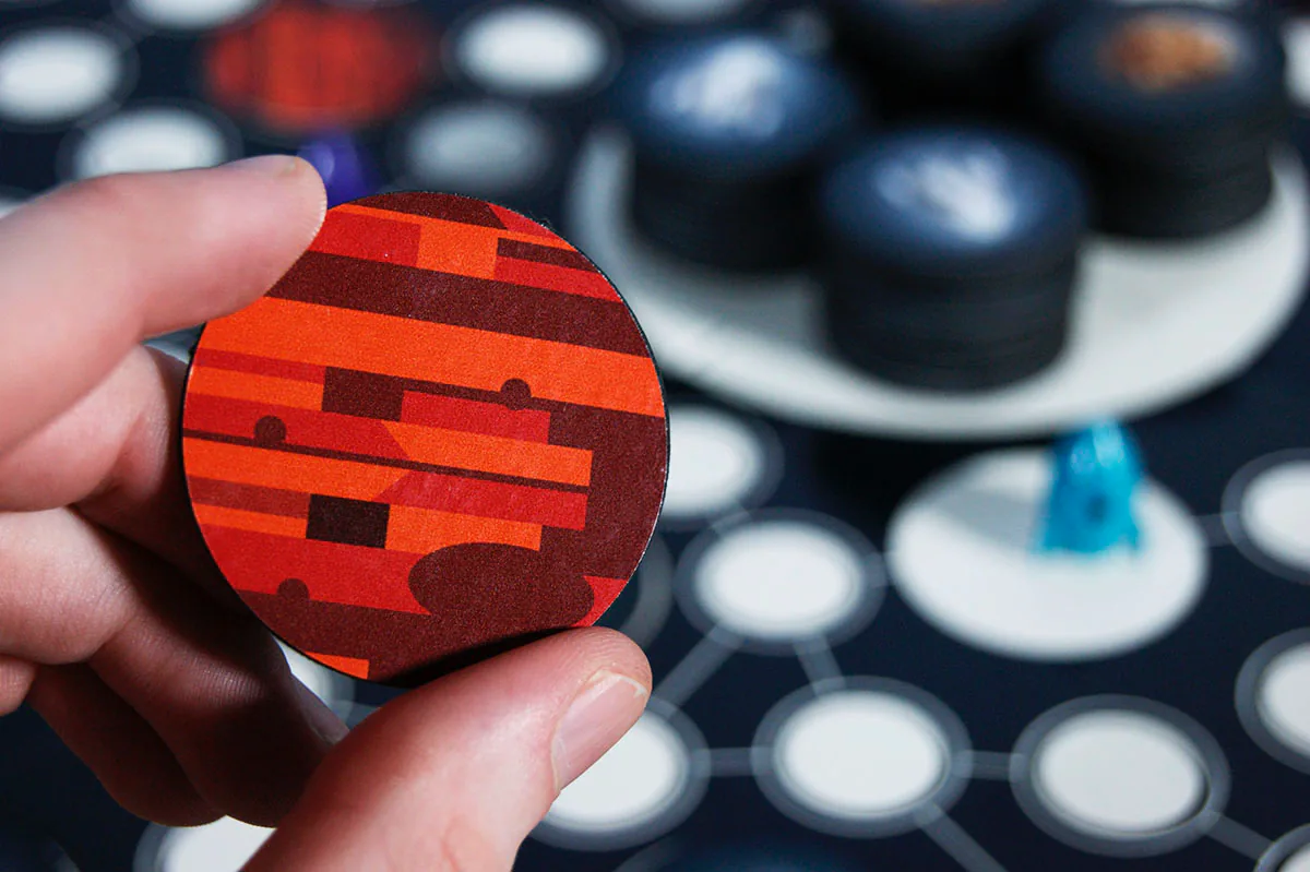 Hand holding a round Terra Nova planet tile printed with a red-and-orange striped pattern